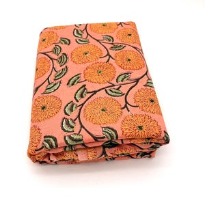 May include: A folded piece of fabric featuring a peach-colored background with a repeating pattern of orange chrysanthemum-like flowers and dark green leaves connected by thin black stems, showcasing a block-printed aesthetic on the textile.