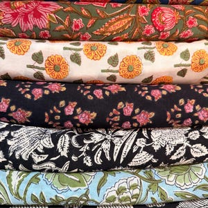 May include: A stack of six folded fabrics featuring various floral and leaf patterns. The designs include blue, olive green, white, black, and light blue backgrounds with printed motifs in shades of pink, orange, green, and white.