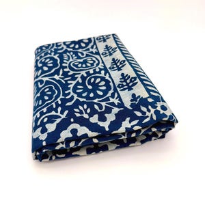 May include: A folded piece of fabric featuring a deep blue background with a white floral and botanical block print pattern. The textile displays a repeating organic motif across the main surface, bordered by a distinct striped edge detail.