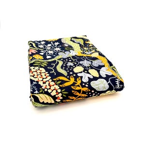 May include: A folded piece of fabric featuring a dark navy blue background with a repeating pattern of stylized botanical elements including yellow, light green, and white flowers, leaves, and winding vines across the surface.