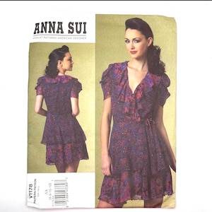 May include: An Anna Sui Vogue Patterns American Designer sewing pattern envelope, number V1178, featuring a short-sleeved dress with a ruffled neckline, a wrap-style bodice, and a tiered skirt, all in a dark purple and blue floral print fabric.