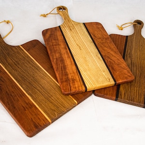 May include: Three wooden chopping boards with different wood grain patterns and colours. Each board has a leather strap handle and a hole for hanging.