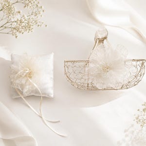 May include: A square white satin ring pillow with a ruffled fabric flower and long ribbons, paired with a small gold wire basket featuring a matching ruffled fabric flower with a pearl center, both accented by delicate white floral sprigs.