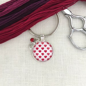 May include: A silver key chain with a red heart patterned charm and a red rhinestone. The key chain is on a white fabric background.