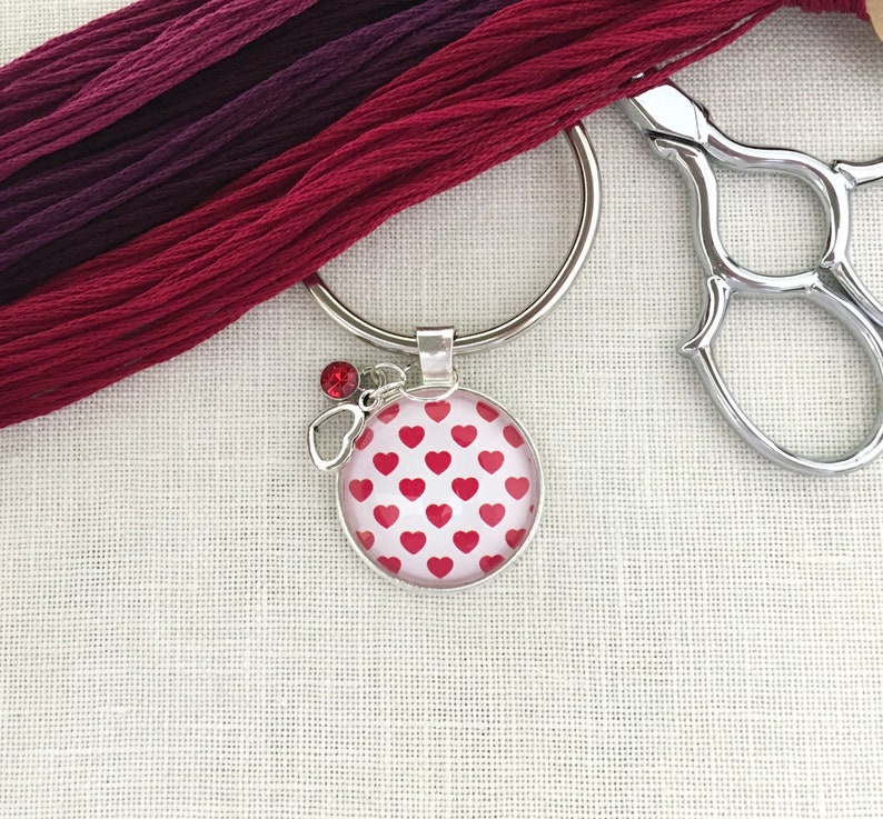 May include: A silver key chain with a red heart patterned charm and a red rhinestone. The key chain is on a white fabric background.