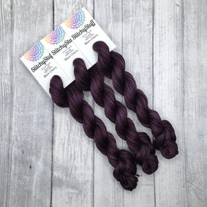 May include: Three skeins of dark purple yarn, labeled "Black Cherry," are displayed on a light gray wooden surface. Each skein is hand-dyed and made of 100% yarn. The labels feature a colorful mandala design and the brand name "Stitchy Stuff."