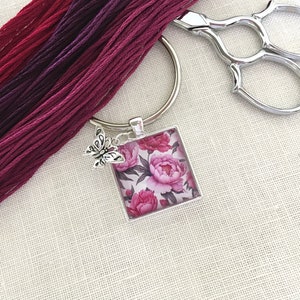 May include: A silver-toned keyring with a square pendant featuring a floral peony design in shades of pink and white. A small butterfly charm dangles from the pendant. Burgundy embroidery floss and silver scissors are also visible.