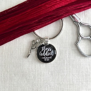 May include: A silver key ring with a black pendant that reads "Floss Addict and proud of it!" in white script. A small pair of scissors charm and red embroidery floss are also visible. A pair of silver scissors is in the background.