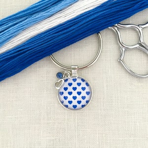May include: A silver key chain with a blue heart pattern charm. The charm is round and has a white background with blue hearts. The key chain is attached to a silver ring.