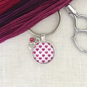 May include: A silver key chain with a pink heart patterned charm and a pink rhinestone. The charm is attached to a silver ring.
