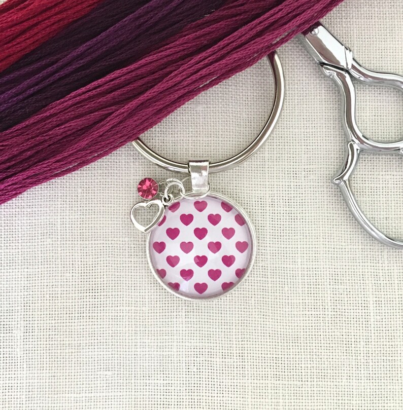 May include: A silver key chain with a pink heart patterned charm and a pink rhinestone. The charm is attached to a silver ring.