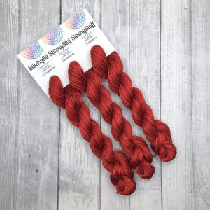 May include: Three skeins of red hand-dyed yarn labeled "Stitchysu Stitchysu Stitchysu" with the text "Hand-Dyed 50 gr 100% cotton Cranberry" on each label.
