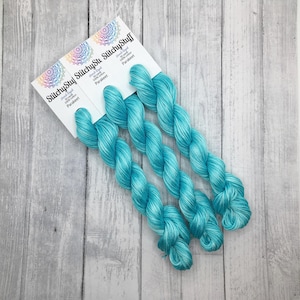 May include: Three skeins of hand-dyed, 100% cotton yarn in a vibrant turquoise color, labeled "Parakeet." The yarn is displayed on a light gray wooden surface, with the "StitchyStuff" logo visible on the packaging.