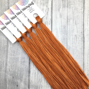 May include: Several bundles of hand-dyed Red Fox embroidery floss from StitchyStuff. Each bundle is tied with a white label that includes the brand name and product details. The floss is a warm, reddish-orange color, and the background is a light gray wood.