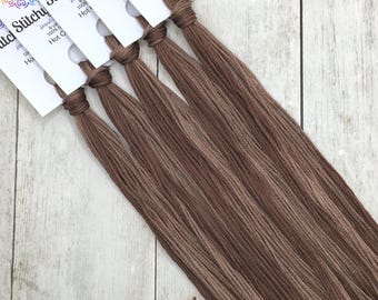 May include: Five skeins of brown embroidery floss, each attached to a white paper tag labeled Stitchy with the text 8 yards, 100% cotton, and Hot Cocoa. The threads are arranged in a fan shape, showcasing the smooth texture of the brown fiber.