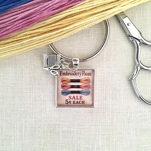 Peut inclure: Un porte-clés argenté avec un pendentif carré portant le texte Embroidery Floss SALE 5¢ EACH et une illustration de trois échevettes de fil. Une petite breloque argentée en forme de bobine de fil et de ciseaux est fixée à l'anneau.