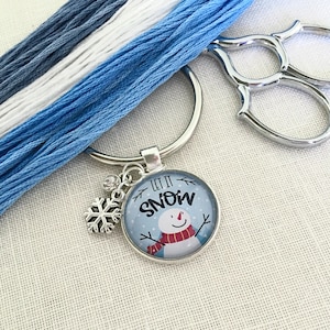 May include: A silver key chain with a blue and white snowman charm that says "Let it Snow". The charm has a snowflake attached to it. The key chain is also attached to a spool of blue and white embroidery floss.