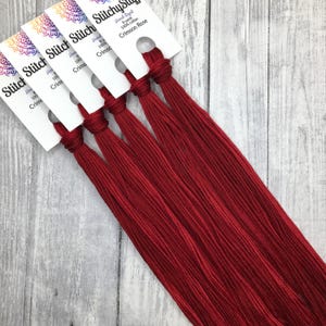 May include: Crimson Rose hand-dyed embroidery floss. The image shows several bundles of red cotton floss, each with a white label. The labels display the brand name and product details. The floss is 8 yards long.