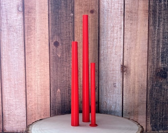May include: Three red cylindrical candles of varying heights, standing on a wooden surface.