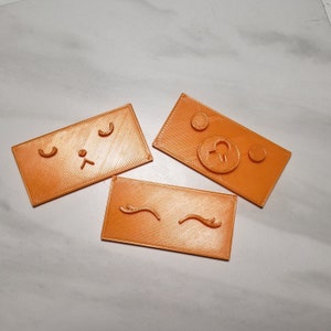 Könnte beinhalten: Drei orangefarbene Stempel aus Kunststoff mit verschiedenen Designs. Der erste Stempel hat ein einfaches Gesicht mit zwei Augen und einer Nase. Der zweite Stempel hat zwei Wimpern. Der dritte Stempel hat einen Kreis mit einem kleinen Kreis darin.