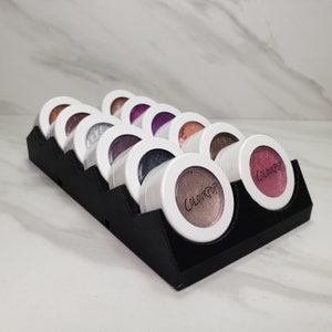 May include: A black eyeshadow palette with 10 round pans of shimmery eyeshadow. The palette is labeled "ColourPop" and is displayed on a black stand.