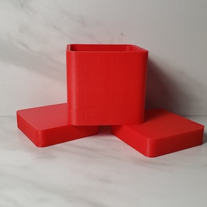 Puede incluir: Un conjunto de tres recipientes rojos brillantes de forma cuadrada. Uno es un cubo, y los otros dos son tapas rectangulares planas. Los objetos están hechos de un material sólido y están colocados sobre una superficie blanca.