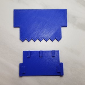 May include: Two blue 3D printed plastic pieces. The top piece has a flat surface and a jagged edge. The bottom piece has a flat surface with three rectangular cutouts.