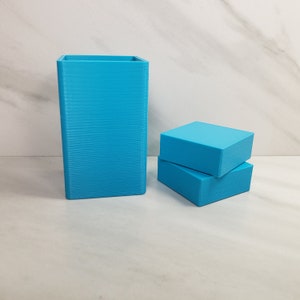 May include: A tall, rectangular, turquoise-colored container stands next to two smaller, turquoise-colored rectangular blocks. The objects are made of a solid material and have a textured surface. The objects are displayed on a white surface.