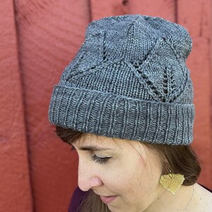 Peut inclure: Un bonnet en tricot gris avec un motif géométrique. Le bonnet est porté par une personne aux cheveux longs bruns et des boucles d'oreilles en feuilles d'or.