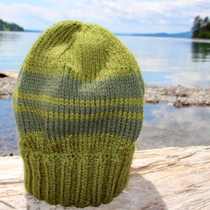 Puede incluir: Un gorro de punto de color verde y gris a rayas con textura acanalada. El gorro está sobre una superficie de madera desgastada.