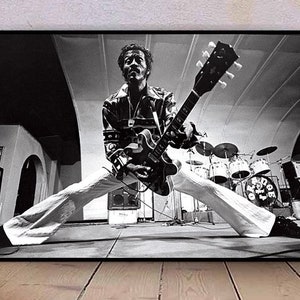 Puede incluir: Fotografía en blanco y negro de un músico tocando una guitarra eléctrica negra con una golpeador blanco. El músico lleva una chaqueta negra y pantalones blancos. La foto está tomada desde un ángulo bajo, y el músico está mirando hacia abajo a la guitarra.