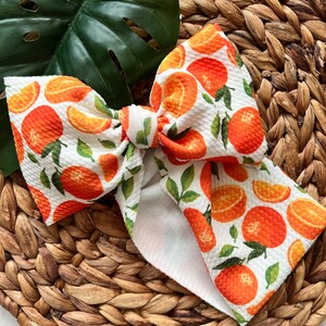 May include: A white fabric bow with a pattern of orange slices and green leaves. The bow is tied in a knot and is lying on a woven brown surface.