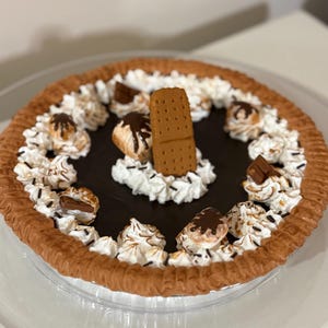 May include: A chocolate pie with a brown crust, topped with white whipped cream, chocolate drizzle, and a graham cracker. The pie is decorated with small chocolate candies and sits on a clear glass plate.