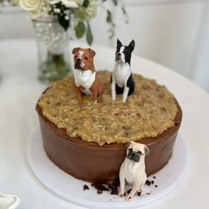 Puede incluir: Un pastel de chocolate con glaseado de chocolate marrón y una cobertura de chocolate alemán blanco y marrón. Tres figuritas de perros están encima del pastel, un perro marrón y blanco, un perro negro y blanco y un perro marrón claro.