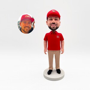 May include: A custom bobblehead figurine of a person wearing a red polo shirt with the logo "Raising Cane's" and a red baseball cap with the same logo. The figurine is standing on a round base.