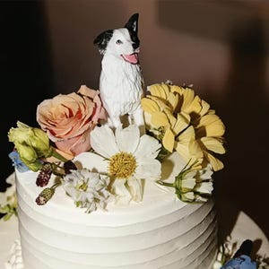 Può includere: Una torta nuziale bianca con una statuetta di cane bianco e nero in cima. La torta è decorata con una varietà di fiori, tra cui rose, margherite e fiori gialli.