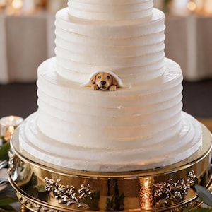May include: A three-tiered white cake with horizontal textured frosting lines. A small, sculpted golden retriever puppy figure is peeking out from under a fold of frosting on the middle tier. The cake sits on a decorative gold metal stand.