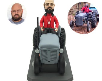 May include: A custom figurine of a person in a red jacket driving a gray tractor. The figurine is on a black base.
