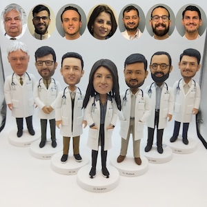 Op de afbeelding: Een verzameling op maat gemaakte bobblehead-beeldjes van medisch personeel, elk gekleed in een witte labjas en een stethoscoop. De beeldjes staan op witte, ronde sokkels, met de namen van de artsen erop. De afbeelding bevat ook portretten van de artsen.