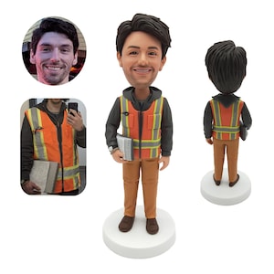 Peut inclure: Une figurine de tête de bobble personnalisée d'une personne portant un sweat à capuche noir, un gilet de sécurité orange et un pantalon marron. La figurine tient un presse-papiers et est debout sur une base blanche.