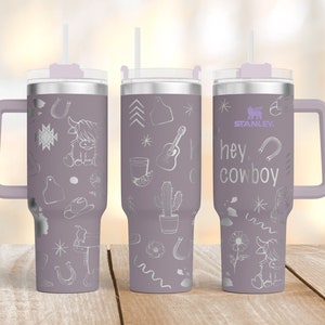 May include: A lavender Stanley tumbler with a handle and straw, featuring etched cowboy-themed designs. The designs include a guitar, cactus, horseshoe, and the text "hey cowboy". The tumbler has a capacity of approximately 1.2 liters.