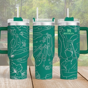 May include: A green Stanley tumbler with a white golf course map design and a white straw. The tumbler has a green handle and lid. The Stanley logo is visible. The design includes a golfer illustration and course hole numbers.