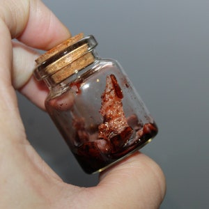 May include: A small glass bottle with a cork stopper, filled with a dark red liquid. The bottle is held in a hand.