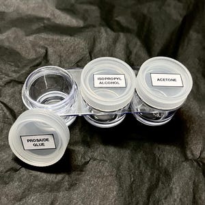 May include: A clear plastic container set with four small, clear jars with white lids. Three jars have labels that read "Isopropyl Alcohol", "Acetone", and "Prosaide Glue". The jars are on a clear plastic tray.
