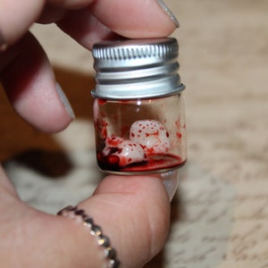 May include: A small glass jar with a silver lid contains a few white, speckled objects in a red liquid. The jar is being held by a hand.