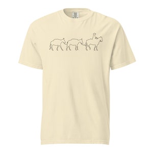 Könnte beinhalten: Ein beiges T-Shirt mit einer schwarzen Linienzeichnung eines Cowboys, der auf einem Pferd reitet und drei Rinder treibt.