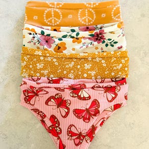 May include: A stack of four pairs of high-waisted underwear. The top pair is mustard yellow with white peace signs. The second is white with pink and orange flowers. The third is mustard yellow with white floral patterns. The bottom is pink with red butterflies.