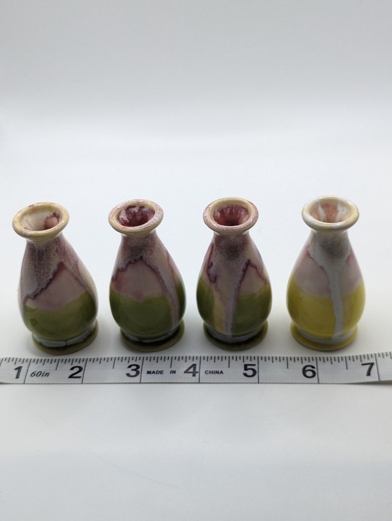 Pu&ograve; includere: Quattro piccoli vasi in ceramica con una smaltatura gialla, verde e rosa. I vasi sono allineati in una fila su una superficie bianca con un metro a nastro.