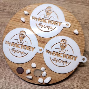May include: Three white plastic stencils with the text "My Factory - Thy Crepes" and a cartoon image of a person wearing a hat and glasses. The stencils are on a wooden surface.