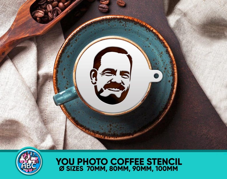 Op de afbeelding: Een turquoise koffiekop en schotel met een sjabloon van een lachend mannengezicht in het koffieschuim. De kop staat op een getextureerd oppervlak met koffiebonen en een houten lepel. De tekst "YOU PHOTO COFFEE STENCIL" is zichtbaar.